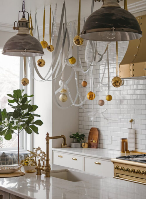 A chic kitchen with industrial-style pendant lights. There are gold ornaments and silver ribbons hanging elegantly, creating a glamorous holiday feel. The kitchen has a marble countertop, a subway tile backsplash, and a gold faucet. There's a potted plant near the window. The floor is made of wood.