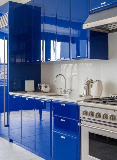 a sleek kitchen with bold blue acrylic cabinets. The cabinets have a glossy finish and are placed above white countertops. There is a stainless steel sink with a faucet next to the cabinets. A stainless steel stove with knobs is positioned on the right. The floor is covered with light grey tiles.