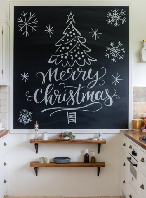 A photo of a white kitchen with a large chalkboard on the wall displaying a hand-drawn Christmas tree, festive "Merry Christmas" lettering, and snowflake doodles. There is a wooden shelf with a few items below the chalkboard. The floor is made of beige tiles.