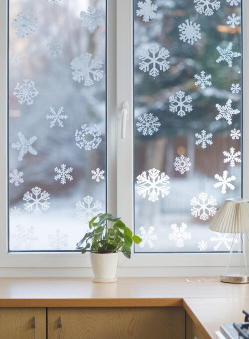 A bright kitchen window covered in snowflake-shaped clings. The window has a snowy outdoor backdrop visible, adding a frosty and festive feel. The kitchen has a wooden cabinet, a white countertop, and a potted plant. There's a lamp on the countertop.