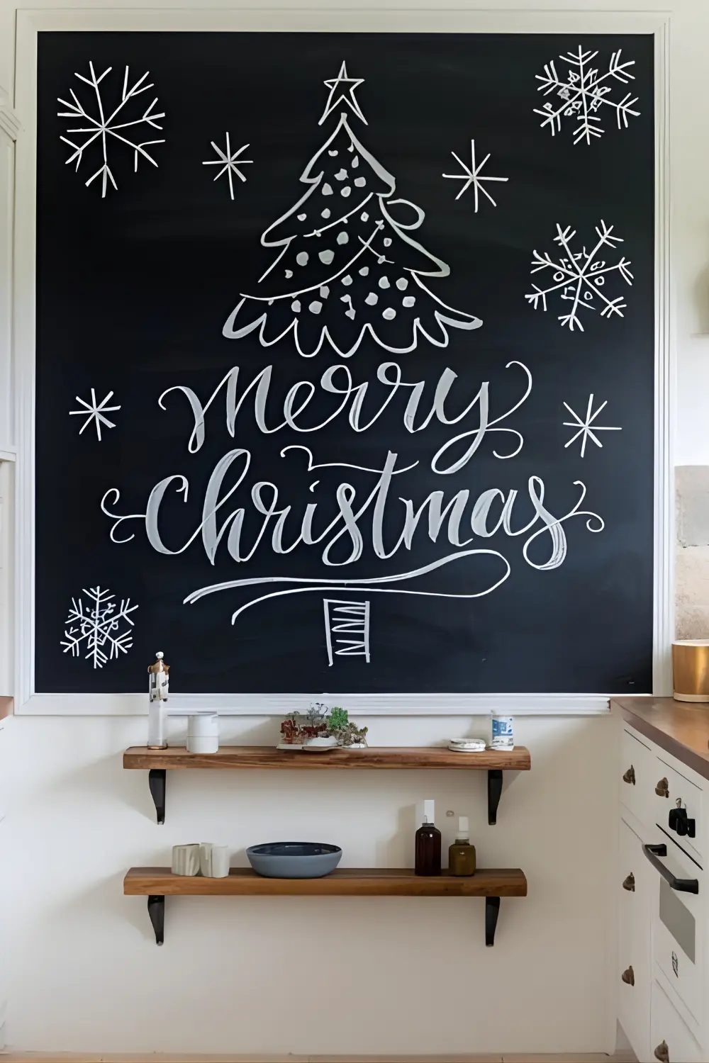 11 best Classic Kitchen Christmas Décor Ideas to Make Your Kitchen Merry and Bright 8 A photo of a white kitchen with a large chalkboard on the wall displaying a hand-drawn Christmas tree, festive "Merry Christmas" lettering, and snowflake doodles. There is a wooden shelf with a few items below the chalkboard. The floor is made of beige tiles.