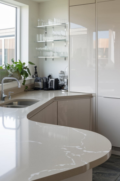 A photo of a modern kitchen with a eased-edged quartz countertop and glossy white cabinets. The countertop is a light grey color with white marks. There is a stainless steel sink with a faucet. There is a glassware storage on the wall, and a plant near the window. The floor is made of dark grey tiles.