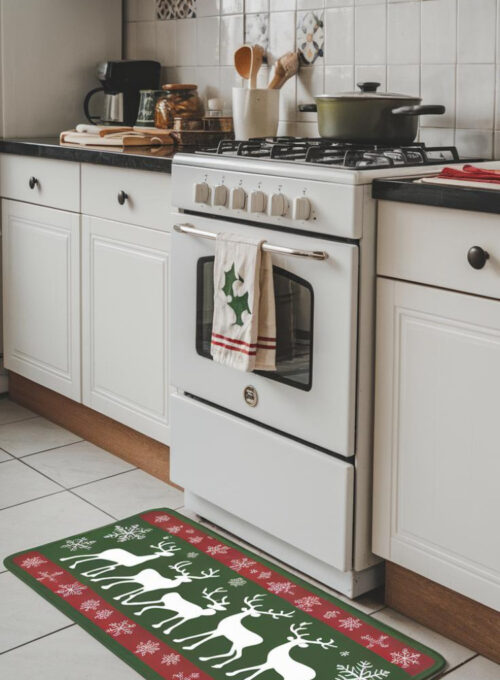 A photo of a modern kitchen with a green and red holiday-themed mat near the stove. The mat features reindeer silhouettes and snowflakes, bringing Christmas cheer to the room. The kitchen has white cabinets, a black countertop, and a white stove. There are also a pot, a wooden spoon, and a dish towel on the counter. The wall behind the stove has a decorative tile pattern.