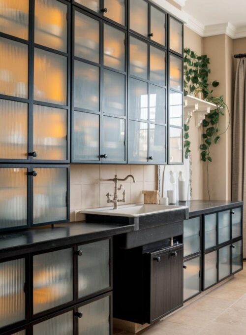 a modern kitchen with frosted glass cabinet doors and black frames. The cabinets are built into a wall and have a sleek, sophisticated design. There's a black countertop with a white sink and faucet. The floor is covered with beige tiles. There's a black cabinet below the sink. The wall has a green plant and a white shelf. The room has a window with curtains.