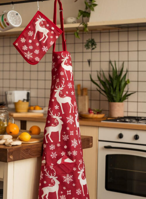 A cozy kitchen scene with a festive red apron with reindeer and snowflakes hanging from a hook. Next to the apron, there are matching oven mitts hanging on a nearby rack. The kitchen has a wooden island with a few ingredients, such as oranges and lemons. There is a plant near the oven. The walls have white cabinets and a tile backsplash. Less
