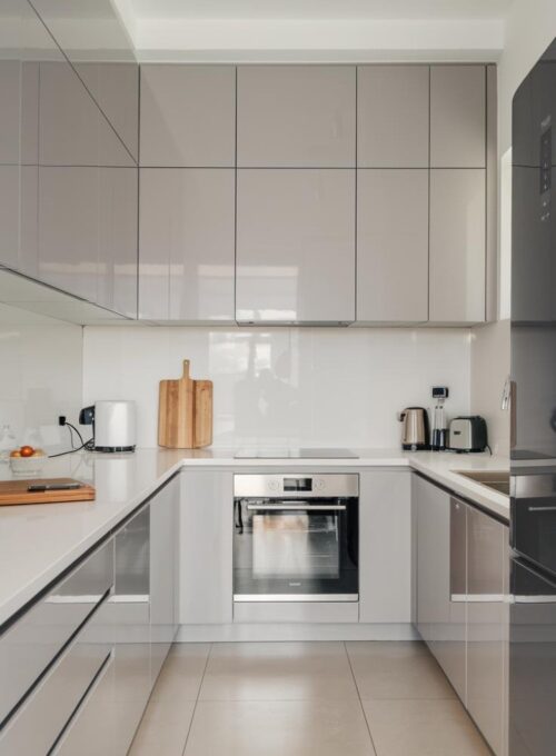 a modern kitchen with sleek grey laminate cabinets, white countertops, and chrome fixtures. The kitchen has a minimalist design with a few appliances, including a refrigerator, oven, and sink. There is a wooden cutting board and a knife on the countertop near the sink. The floor is tiled.