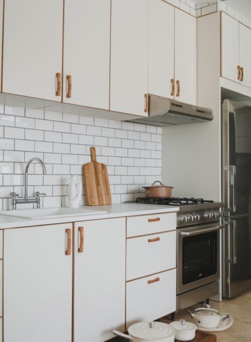 A minimalist kitchen with white melamine cabinets with a matte finish, stainless steel appliances, and a subway tile backsplash. The cabinets have wooden handles. There is a white sink with a stainless steel faucet. A stainless steel oven and a refrigerator are placed against the wall. There is a wooden cutting board on the counter. A set of white pots and pans are placed on the floor. The floor is made of beige tiles.