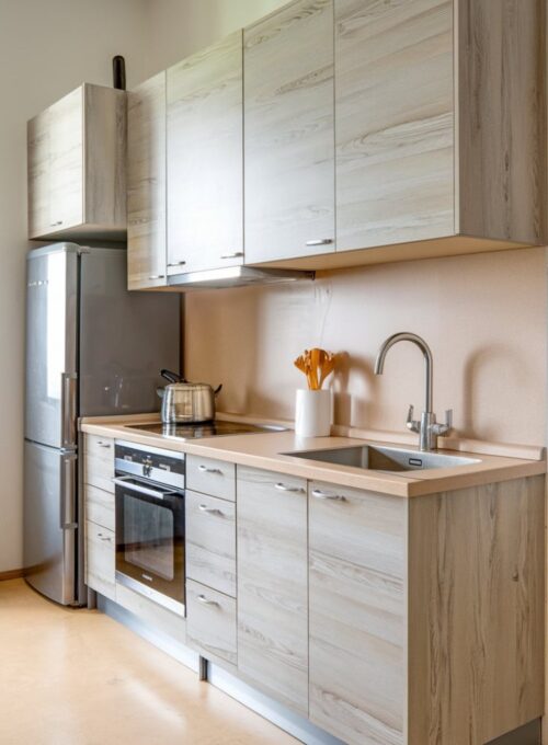 a budget-friendly kitchen with particleboard cabinets featuring a light grey woodgrain veneer. The cabinets have a simple design and are installed above a beige countertop. There is a stainless steel sink mounted on the countertop, next to which is a stainless steel faucet. A stainless steel refrigerator and a stove are placed at the back of the kitchen. There is a beige floor and a white wall in the kitchen.