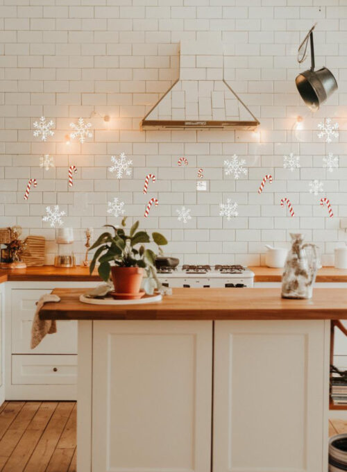 A photo of a bright white kitchen with a subway tile backsplash. There are temporary stickers of snowflakes and candy canes on the tiles, adding a whimsical Christmas touch. The kitchen has a large island in the center with a wooden countertop and a few items on it, including a potted plant and a decorative object. There is a pot hanging above the island. The floor is made of wooden planks. The lighting is warm.