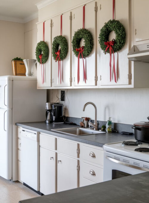 A photo of a modern kitchen with white cabinets. There are small green wreaths adorned with red ribbons hung on each cabinet door. The countertops are a dark grey material. There is a stainless steel sink with a faucet in the middle of the room. A white fridge is placed against the wall on the left. There is a black pot on the stove. The floor is covered with beige tiles.