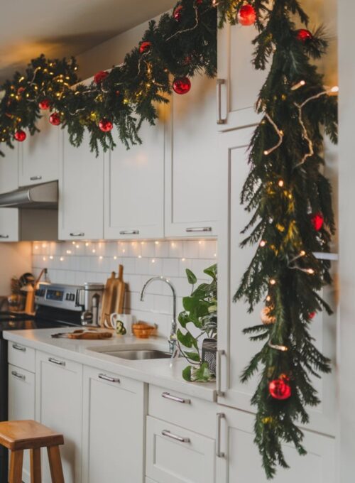 Classic Kitchen Christmas Décor Ideas, A modern kitchen with white cabinets, a lush pine garland draped across the top, with fairy lights and red ornaments woven in, creating a warm and festive ambiance. There's a wooden stool near the sink. A green plant is placed on the counter.