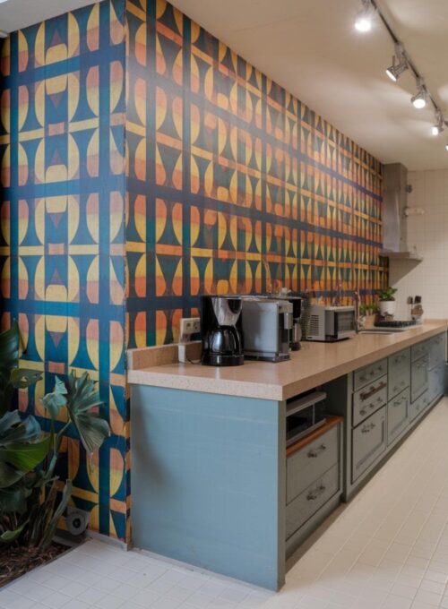 A photo of a contemporary kitchen with a wallpapered accent wall featuring large, colorful geometric patterns. The patterns are a mix of blue, orange, and yellow. The wall is adjacent to the kitchen counter, which has a beige surface and several appliances. The floor is covered with white tiles. There is a potted plant near the wall. The lighting is bright.