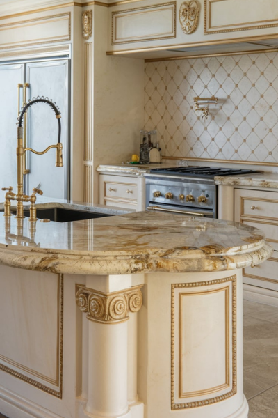 A photo of a luxury kitchen with a double ogee-edged granite countertop. The countertop has two stacked curves and a grand, elaborate design. There is a sink on the left, and a gold faucet with a curved spout. There are also a few appliances, such as a refrigerator, oven, and dishwasher. The backsplash has a unique pattern and is adorned with gold accents. The floor is made of tiles.