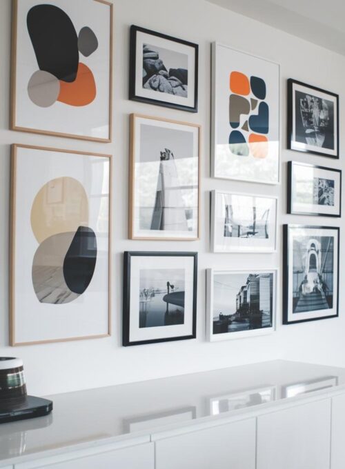 A photo of a modern gallery wall with abstract art and framed black-and-white photos. The wall has a white background and is above a sleek white countertop. The abstract art pieces have various shapes, sizes, and colors. The black-and-white photos are of different subjects, including nature, architecture, and people. The gallery wall is arranged in a way that creates a visually pleasing layout.