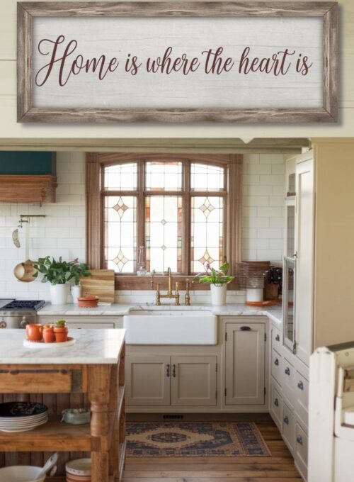 A cozy farmhouse kitchen with a distressed wooden frame holding an inspirational quote in a script font. The kitchen has a vintage feel, with a wooden island, beige cabinets, and a marble counter. The quote reads "Home is where the heart is." There are potted plants and a few cooking utensils on the counter. The floor is made of wood, and there is a patterned rug in the middle. The background is a window with a wooden frame and stained glass.