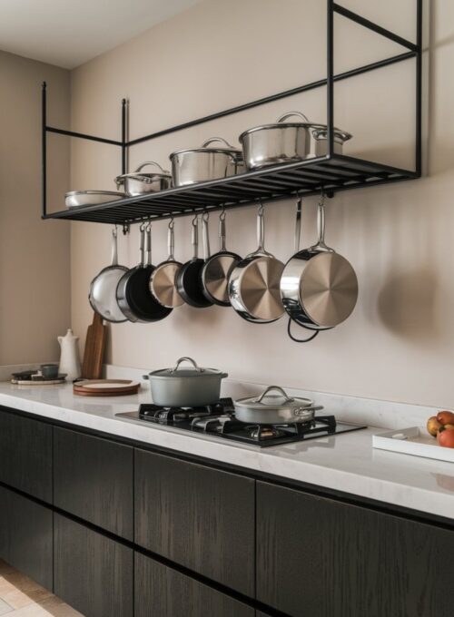 A modern kitchen with a sleek black metal pot rack hanging above a white marble countertop. There are stainless steel pots and pans hanging neatly from the rack. The pots and pans are placed on a white ceramic plate. The wall behind the rack is painted beige. The floor is covered with beige tiles.