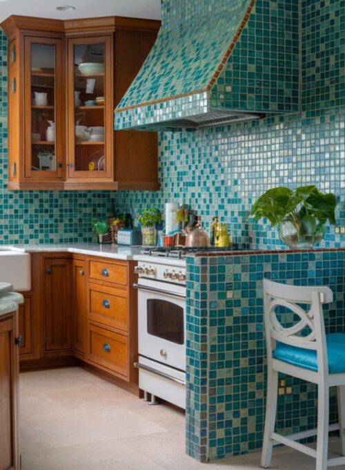 A photo of a vibrant kitchen with a mosaic tile backsplash in shades of blue and green. The backsplash makes a bold statement behind the stove and extends to the dining area. There are wooden cabinets, a white countertop, and a white stove. A green plant sits on the countertop. A white chair with a blue cushion is placed near the dining area. The floor is covered with beige tiles.