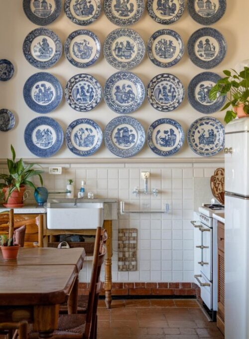 A photo of a traditional kitchen with a symmetrical arrangement of blue and white decorative plates hanging on the wall. The plates have a classic blue and white pattern. There is a wooden table in the kitchen with a few chairs. The kitchen has a white sink, a stove, and a refrigerator. There are also a few potted plants in the kitchen. The floor is made of tiles.