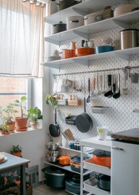 How to Organize a Small Kitchen Without Cabinets, a compact kitchen with a white pegboard wall. The pegboard is neatly filled with pots, pans, and cooking utensils, organized in a way that maximizes space and makes everything easily accessible. There's a window with a curtain, a few potted plants, and a few hanging items. The floor is tiled, and there's a small table near the window.