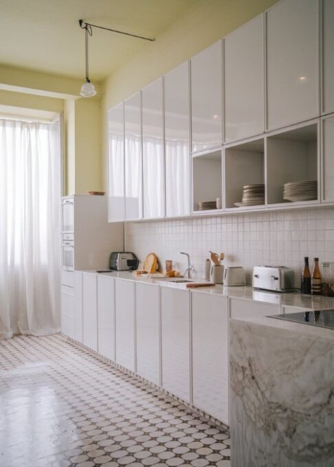 Kitchen Cabinet Colors for White Tile Floors, A minimalist, airy kitchen with a bright color scheme. The room has sleek white cabinets with a few dishes inside. The floor is made up of patterned white tiles. A marble countertop is positioned next to the cabinets. A few items, including a toaster, a kettle, and a few bottles, are placed on the countertop. The room has a window with white curtains and a light fixture hanging from the ceiling.