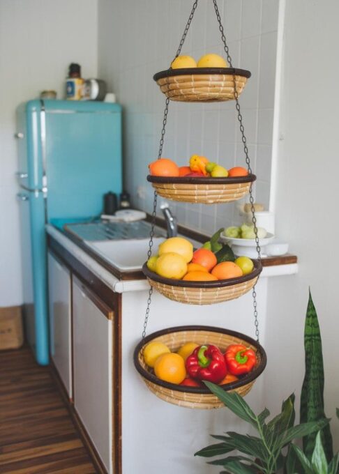 a small kitchen with a three-tiered hanging basket. The basket is filled with colorful fruits and vegetables, including lemons, oranges, and red bell peppers. The basket is hung above the kitchen counter. The kitchen has a white sink, a blue fridge, and a wooden floor. A green plant is placed near the basket.