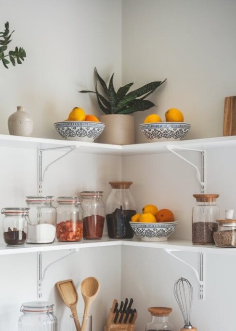 a minimalist kitchen with white corner shelves filled with glass jars, decorative bowls, and small kitchen tools. The jars contain various ingredients, such as spices, dried fruits, and nuts. The bowls are placed on top of the jars and contain fresh fruits, like lemons and oranges. The kitchen tools include a wooden spoon, a spatula, and a whisk. The background is a white wall with a few decorative items, such as a plant and a small vase.