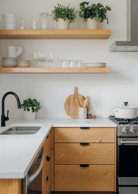 a modern kitchen with minimalistic wooden floating shelves. The shelves hold neatly arranged kitchenware, glassware, and potted herbs for added décor. The kitchen has a white countertop and backsplash, a stainless steel sink, and a black faucet. There's a white pot on the stove. The floor is made of gray tiles.