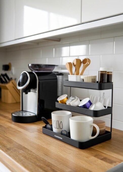 a modern kitchen with a sleek black countertop organizer filled with coffee pods, mugs, and stirrers, creating a stylish and compact coffee station. The organizer is placed on a wooden countertop. There's a coffee machine beside the organizer. The kitchen has white cabinets and a backsplash.