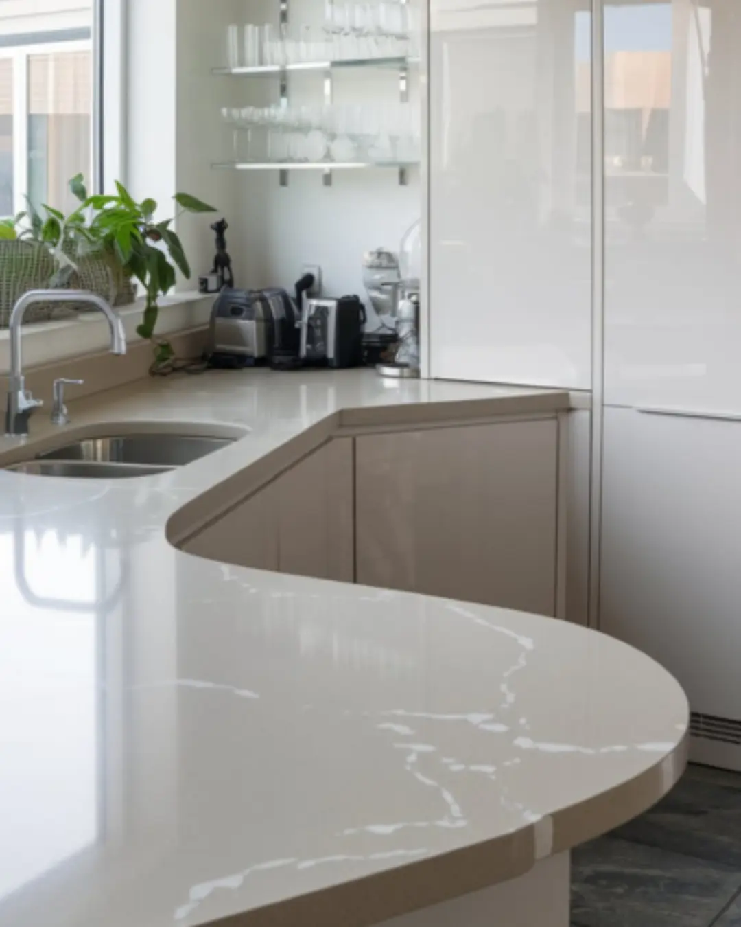A photo of a modern kitchen with a eased-edged quartz countertop and glossy white cabinets. The countertop is a light grey color with white marks. There is a stainless steel sink with a faucet. There is a glassware storage on the wall, and a plant near the window. The floor is made of dark grey tiles.