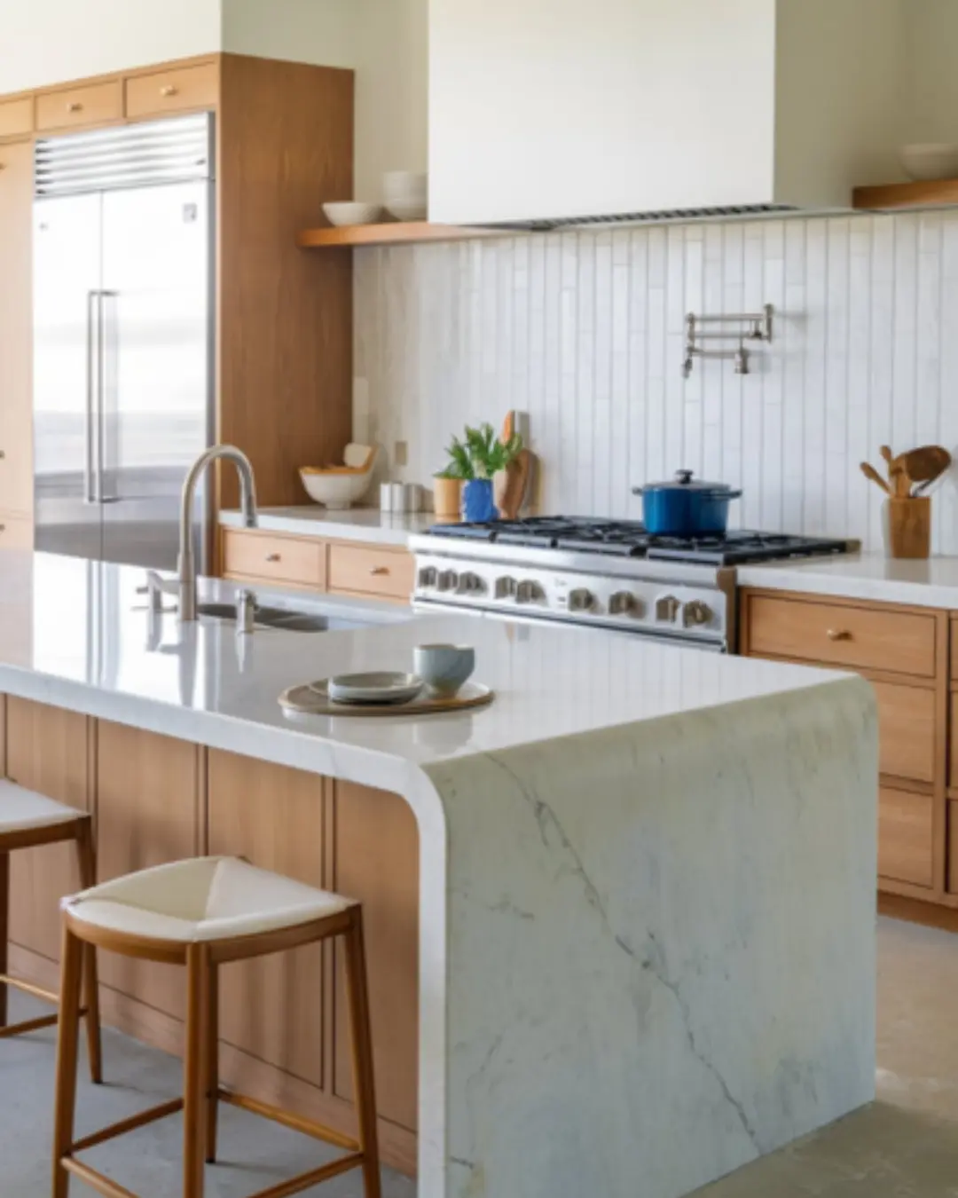 A photo of a bright, open kitchen with a half-bullnose-edged countertop. The countertop is made of marble stone and gently curves on top and flat underneath for a modern twist. The kitchen has stainless steel appliances, a white backsplash, and wooden cabinets. There is a pot on the stove and a dish in the sink.