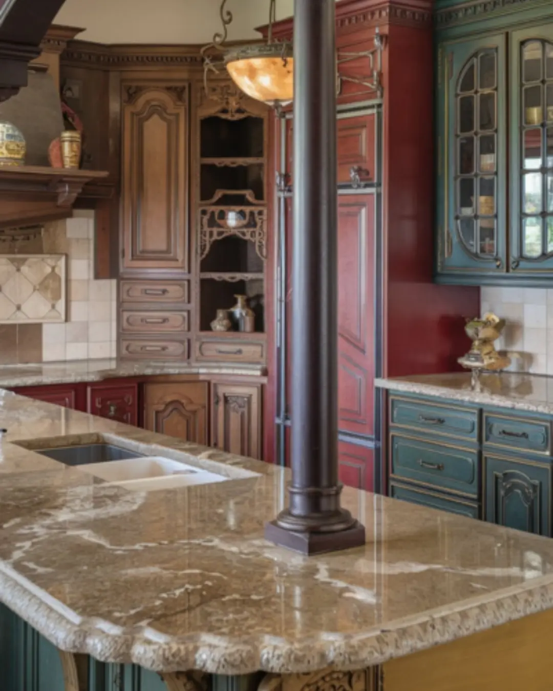 A photo of a traditional kitchen with an ogee-edged granite countertop. The countertop extends into a small island in the middle of the room, with a dark wooden post supporting an arched beam. The cabinets are made of ornate wood and have intricate detailing, with some featuring curved edges and others having decorative carvings. The cabinets vary in color, with some being a deep red and others a dark green. The room has a refined look, with a vintage chandelier hanging above the island and a few decorative items placed on the countertops. Less
