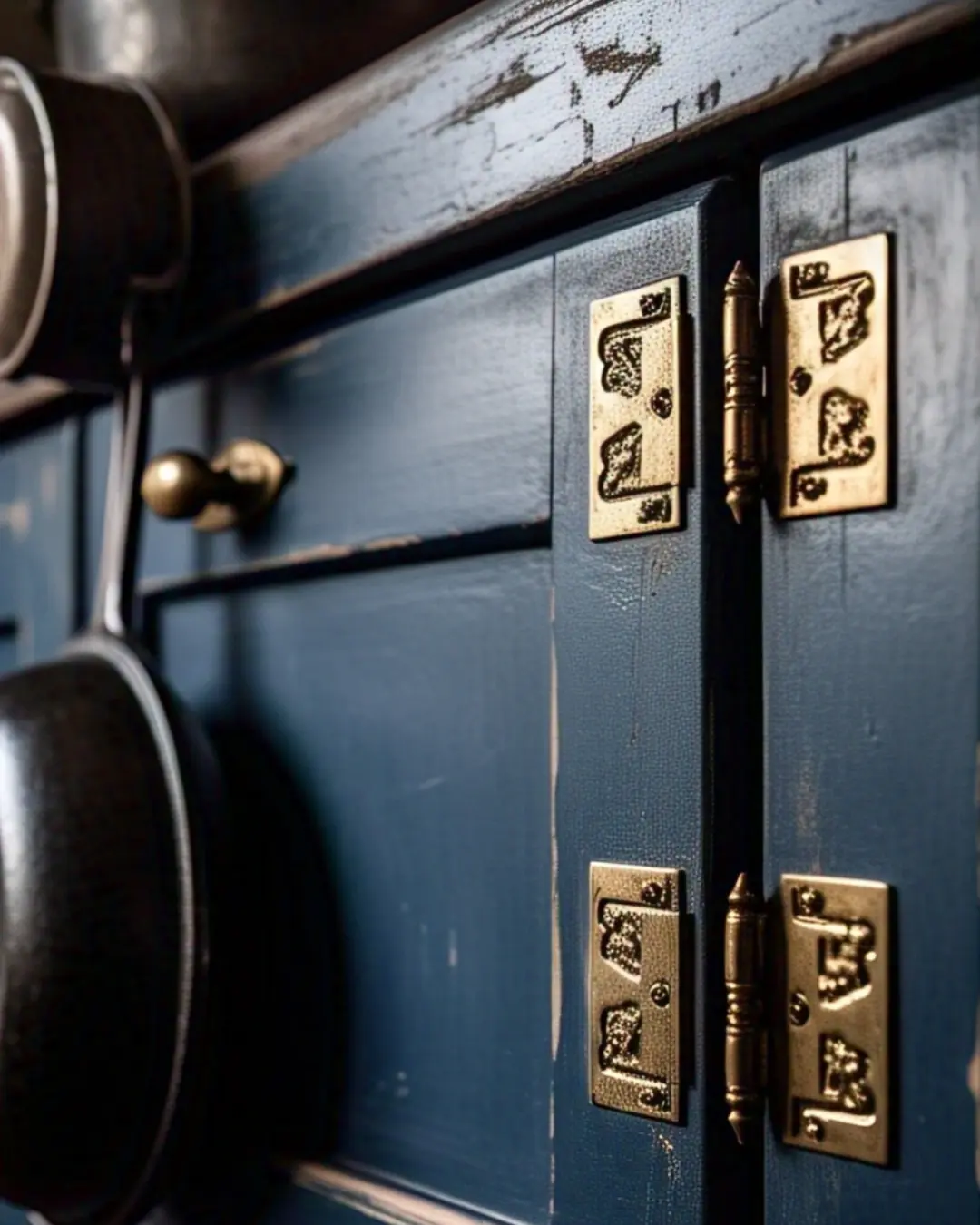 Close-up of distressed navy blue cabinets with gleaming brass surface hinges, their patina telling years of kitchen stories. Morning light catches the hinge's decorative screws while cast iron pans hang nearby, whispering "this kitchen means business."